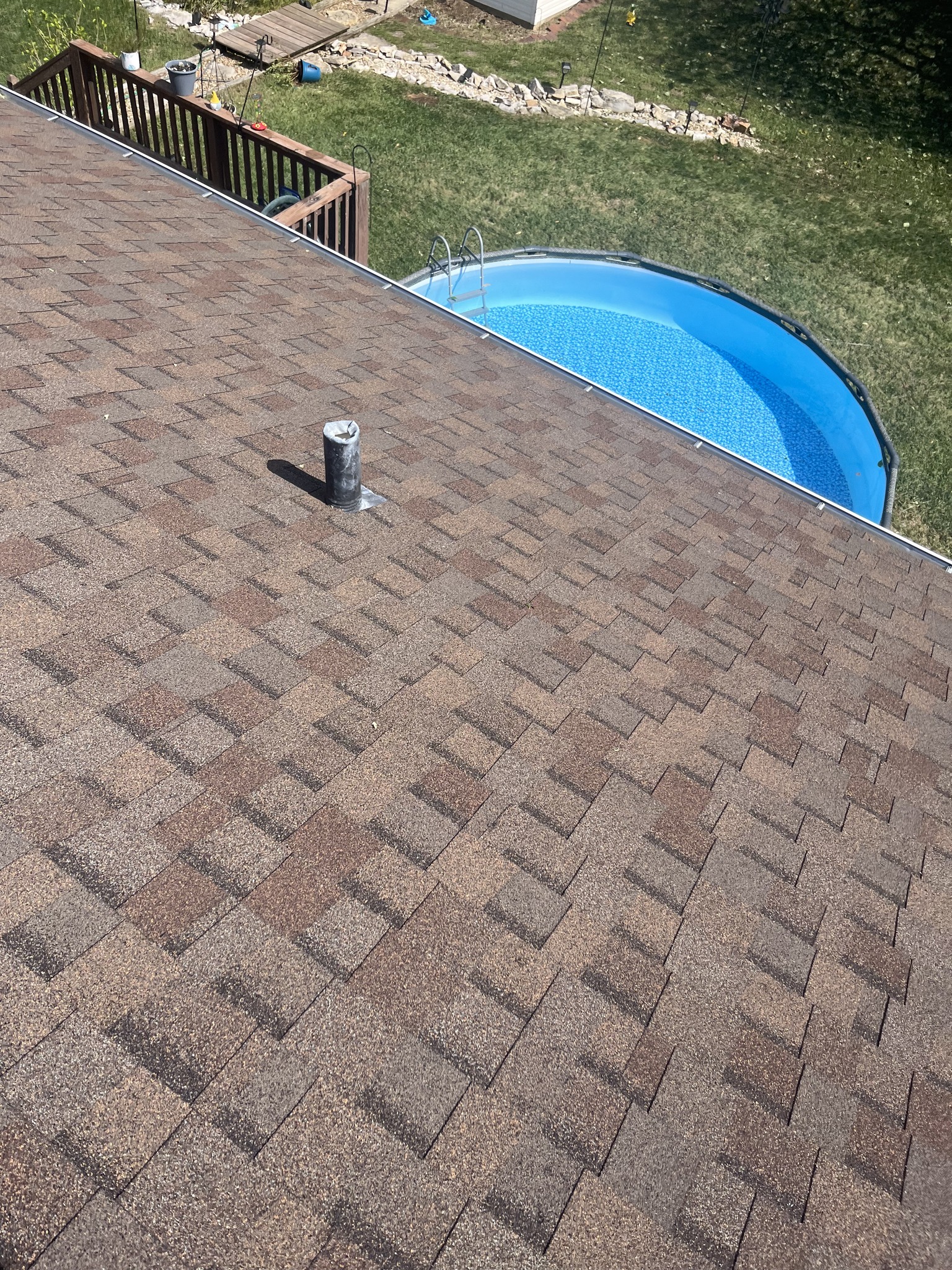 View from a roof with brown shingles, featuring a vent pipe; a round above-ground pool and a wooden deck are visible in the backyard below.