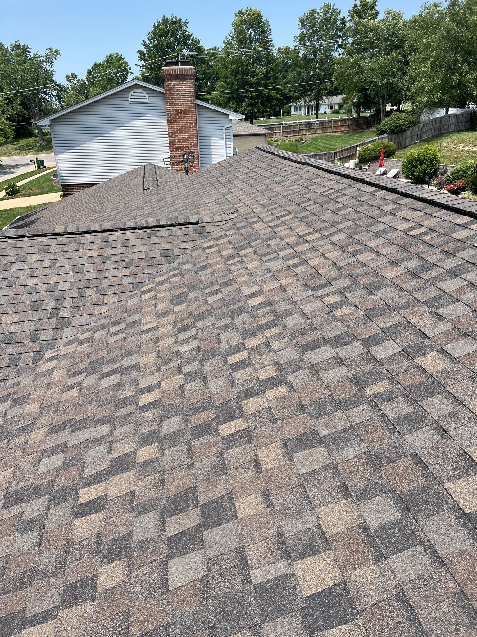 A close-up view of a residential roof with brown asphalt shingles, looking toward a brick chimney and a neighboring house, with trees and lawns in the background.