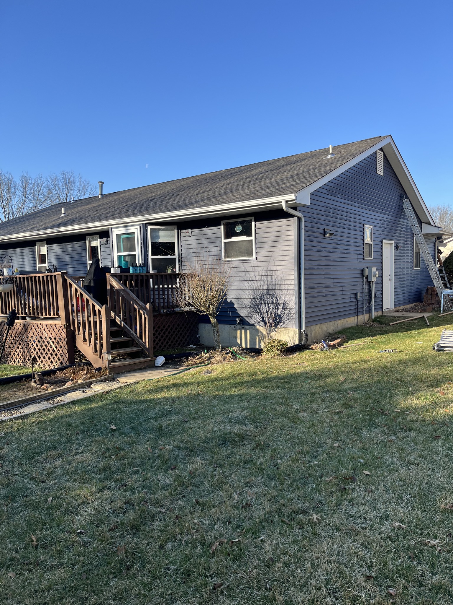 Single-story gray house with white trim, wooden deck, and steps leading to the backyard, on a clear day with blue sky and green lawn.