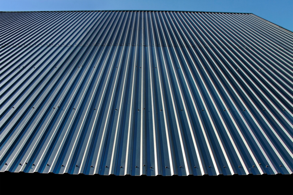A close-up view of a corrugated metal wall or roof, photographed from below, with clear blue sky visible at the top.