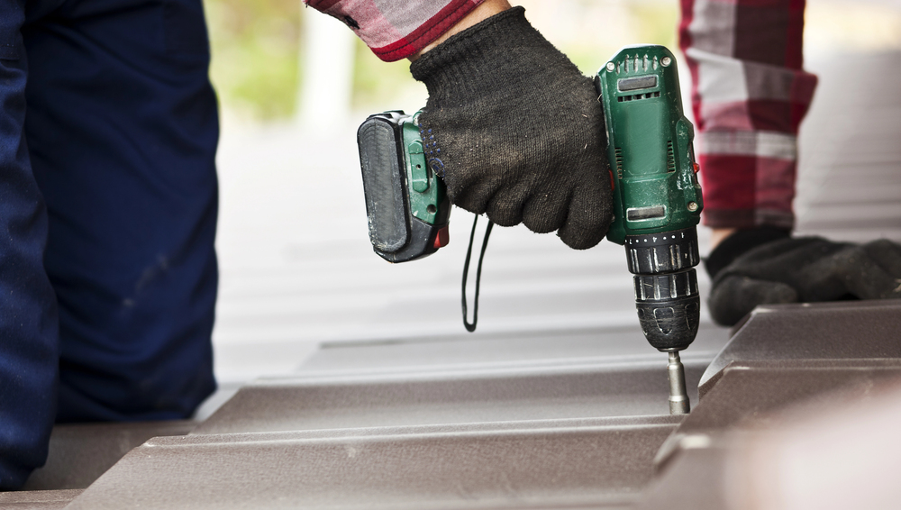 A worker wearing gloves uses a cordless power drill to install or fasten metal roofing sheets.