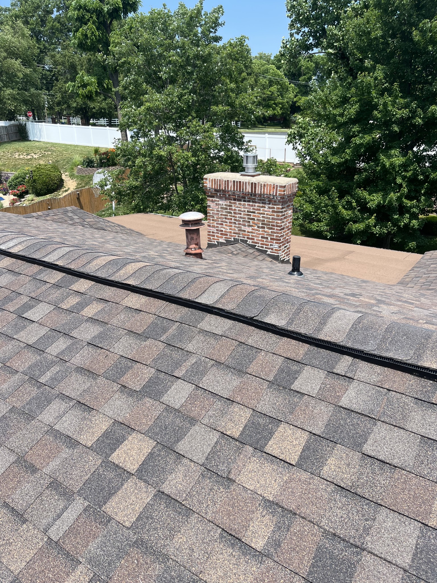 View of a rooftop with brown asphalt shingles, a brick chimney, two metal vents, and surrounding green trees under a clear sky.