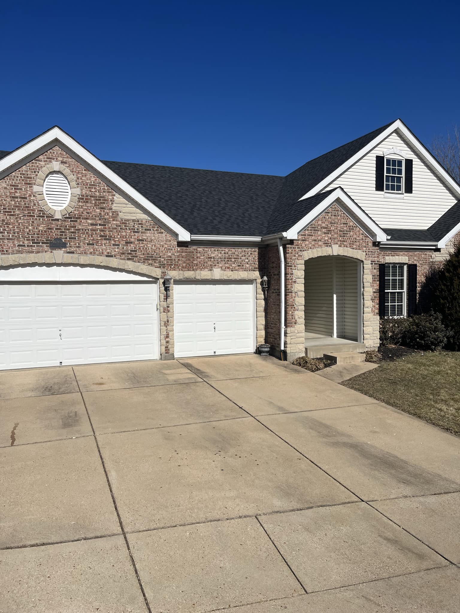 A suburban brick house with a three-car garage, arched entryway, and a concrete driveway under a clear blue sky.