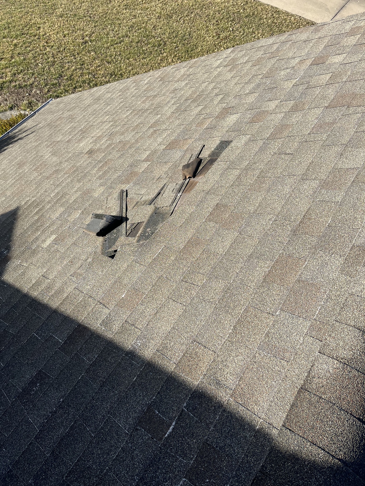 Several roof shingles are missing or damaged, exposing underlying materials and boards, while the rest of the roof appears intact. Grass and pavement are visible in the background.